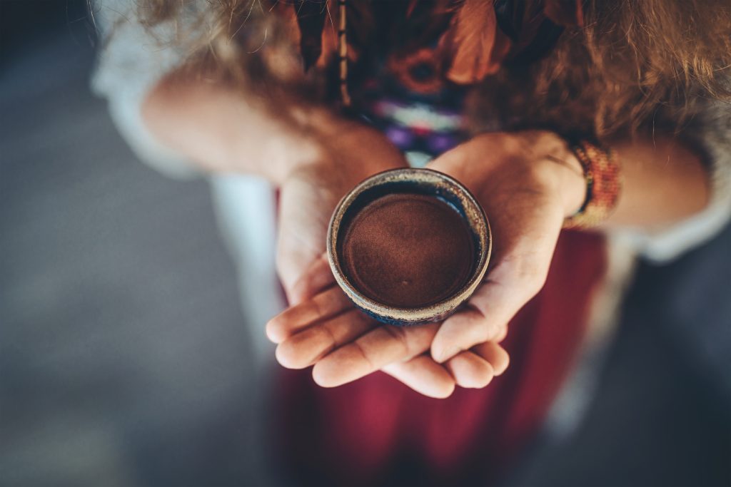 Hands holding a small, round bowl filled with a brown substance, surrounded by colorful fabric and warm lighting.