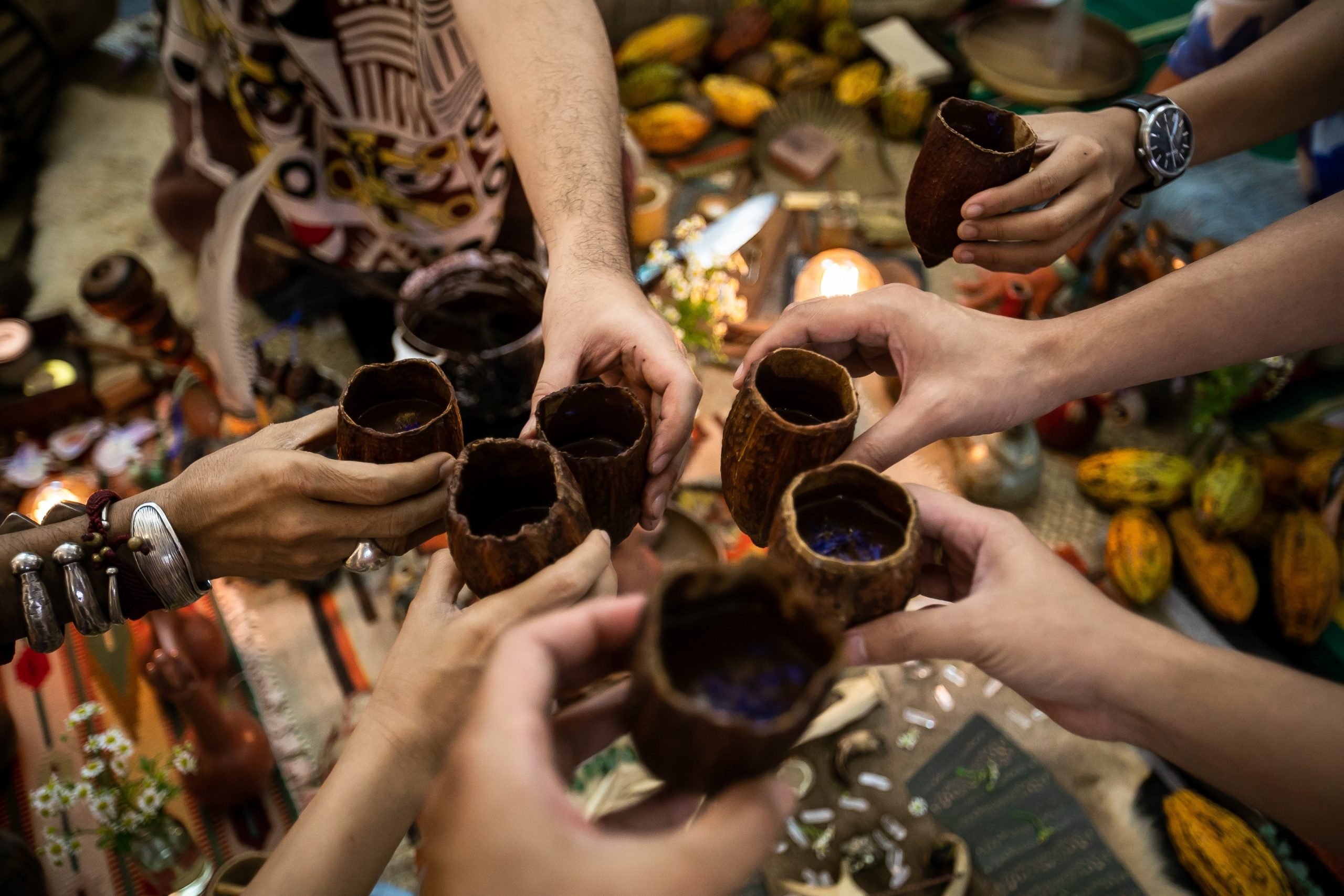 Hands holding traditional cups in a circle, surrounded by colorful cacao pods and decorative items on a table.