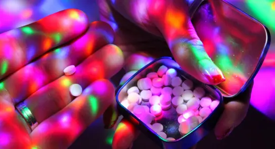 Hands holding white pills, with a colorful, vibrant light pattern reflecting off surfaces.