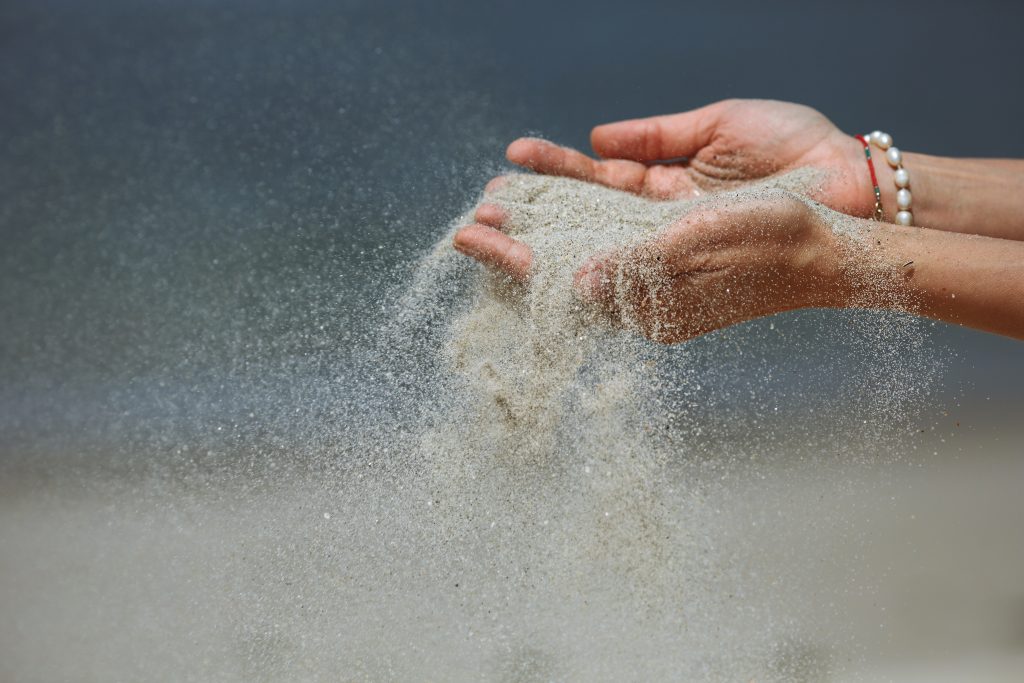 Hands releasing fine sand, creating a shimmering cloud against a blurred beach background.