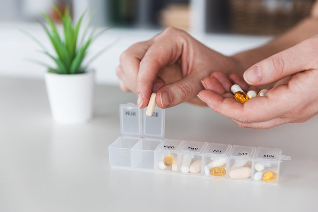 Hands sorting pills from a weekly pill organizer, with a small plant in the background.