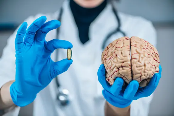 A healthcare professional in blue gloves holds a capsule in one hand and a model brain in the other.