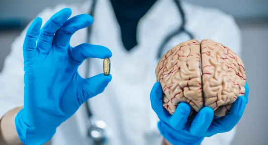 A healthcare professional in blue gloves holds a capsule in one hand and a model brain in the other.