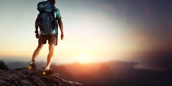 A hiker stands on a rocky outcrop, gazing at a vibrant sunset over distant mountains.