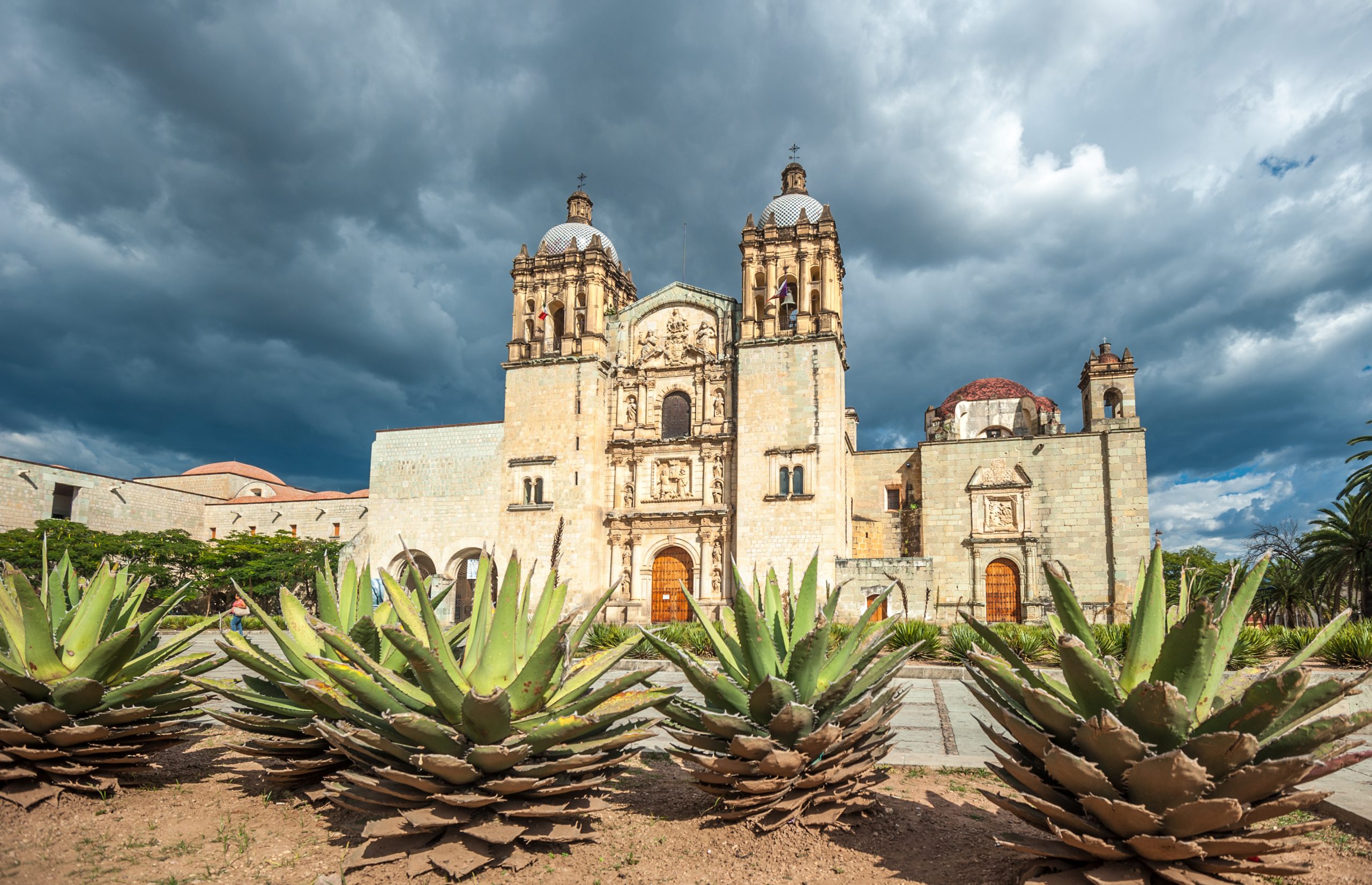 Historic church with ornate architecture, surrounded by agave plants under a dramatic, cloudy sky.