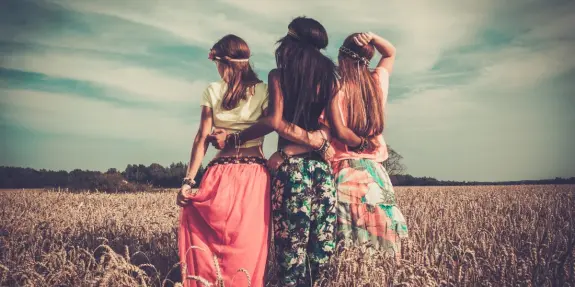 Three women stand together in a wheat field, wearing colorful skirts and floral accessories, gazing at the horizon.