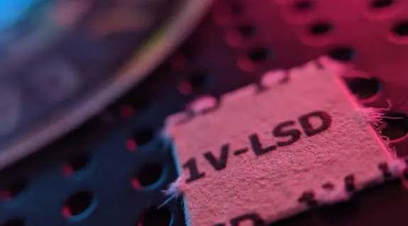 Close-up of a small square blotter paper labeled "1V-LSD" on a textured surface with colorful lighting.