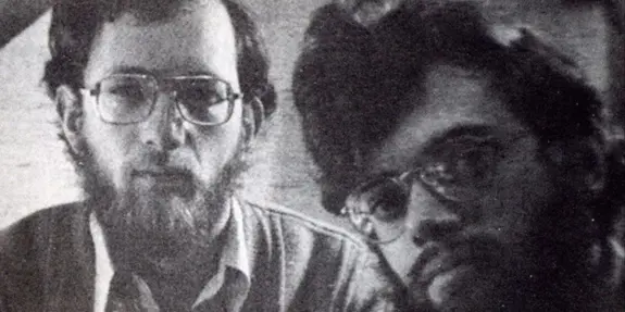 Two men with glasses and beards, looking directly at the camera, set against a blurred background.