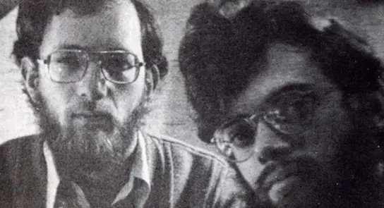 Two men with glasses and beards, looking directly at the camera, set against a blurred background.