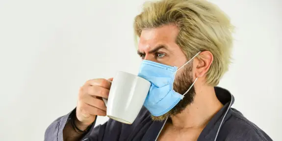 Man with blonde hair wearing a blue mask sips from a white mug, dressed in a robe against a plain background.