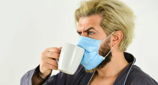 Man with blonde hair wearing a blue mask sips from a white mug, dressed in a robe against a plain background.