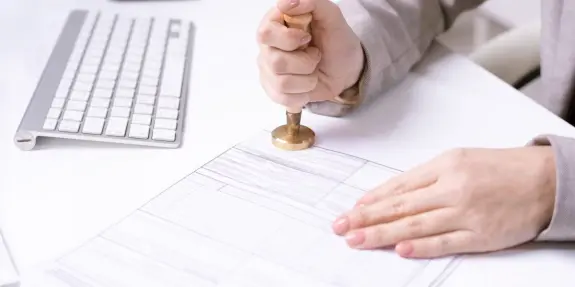 A hand pressing a brass stamp onto a document, with a computer keyboard in the background.