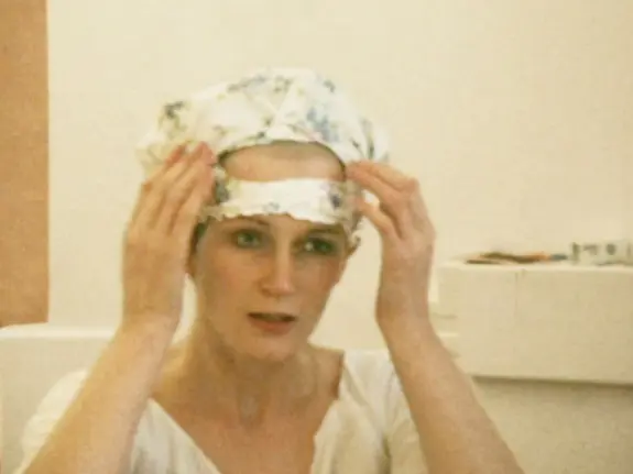 Woman adjusting a floral headscarf, looking thoughtfully at her reflection in a light-colored room.