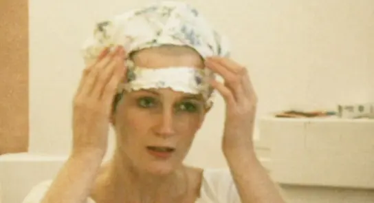 Woman adjusting a floral headscarf, looking thoughtfully at her reflection in a light-colored room.