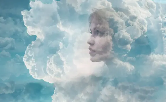 A serene profile of a young woman blends with soft, billowing clouds against a blue sky.