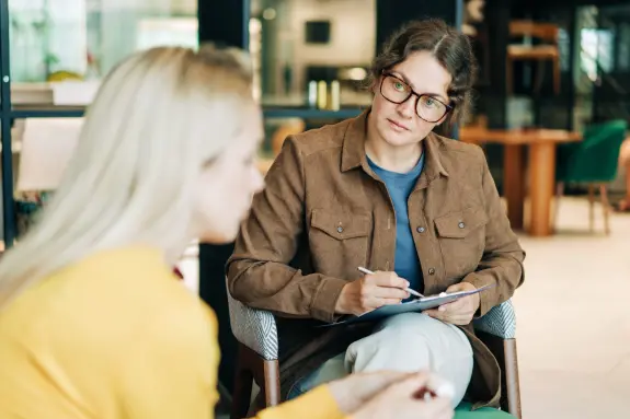 A woman in glasses attentively listens to another woman, taking notes in a modern, well-lit setting.