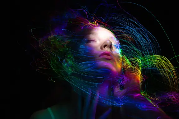 A woman with closed eyes surrounded by vibrant, swirling colorful light trails against a dark background.