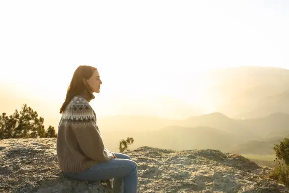 A woman sits on a rock, gazing at a sunlit mountain landscape, wearing a cozy sweater.