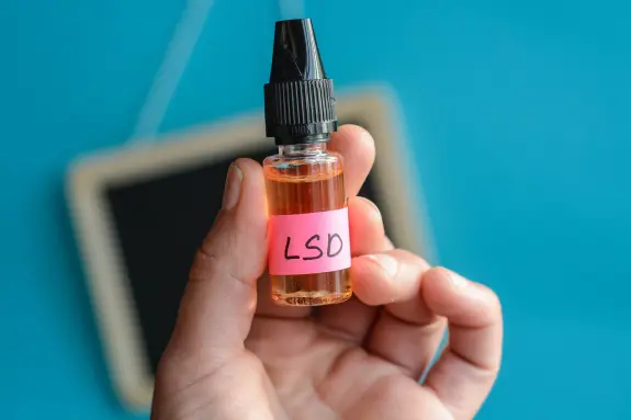 A hand holds a small bottle labeled "LSD" with a bright pink sticker against a blue background.
