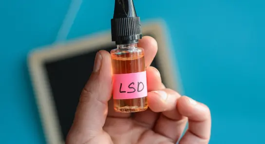 A hand holds a small bottle labeled "LSD" with a bright pink sticker against a blue background.