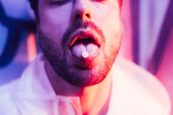 Close-up of a man with a pill on his tongue, illuminated by vibrant pink and purple lights.