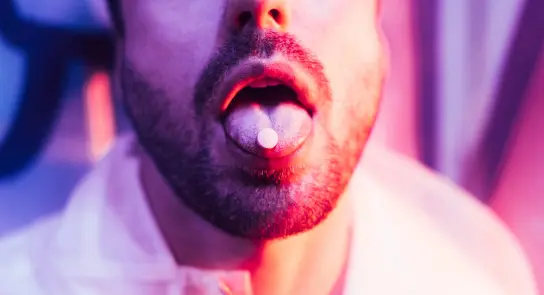 Close-up of a man with a pill on his tongue, illuminated by vibrant pink and purple lights.
