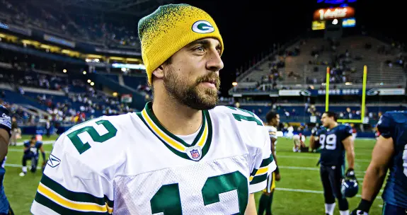 A man in a Green Bay Packers jersey and yellow beanie smiles on the field after a game, stadium lights in the background.
