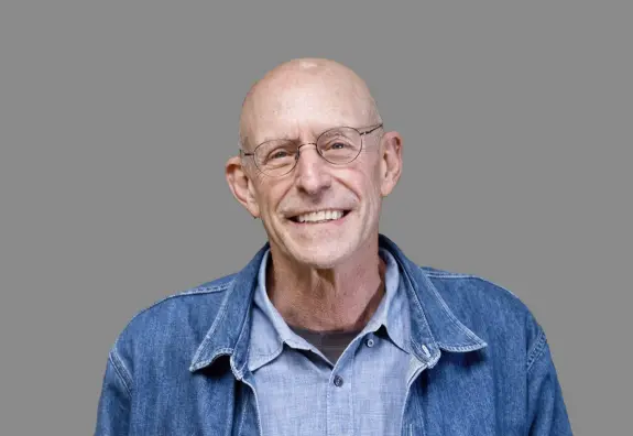 Smiling bald man with glasses, wearing a denim jacket over a light shirt, against a gray background.