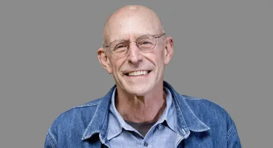 Smiling bald man with glasses, wearing a denim jacket over a light shirt, against a gray background.