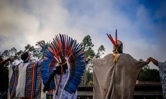 Two individuals in traditional attire, one with a colorful feathered headdress, stand with arms raised against a cloudy sky.