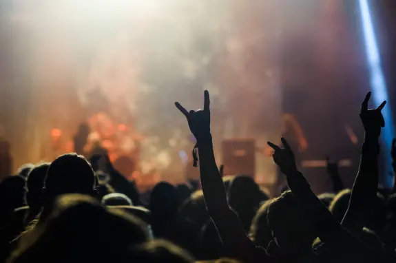 Crowd at a concert with raised hands making rock gestures, illuminated by stage lights.