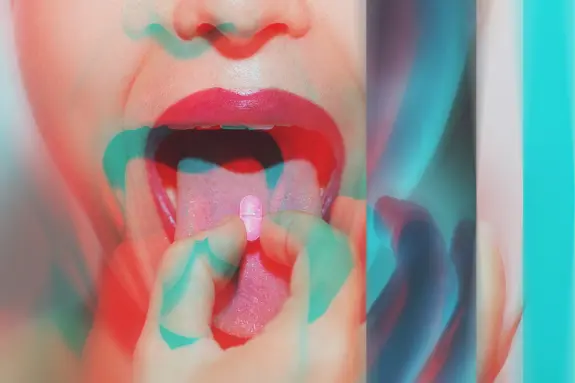 A close-up of a mouth with a hand holding a pink pill, surrounded by vibrant, abstract colors.