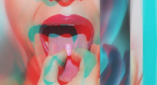 A close-up of a mouth with a hand holding a pink pill, surrounded by vibrant, abstract colors.
