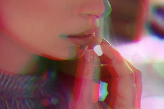 A close-up of a woman’s lips as she holds a small pill, with a colorful, blurred effect surrounding her face.