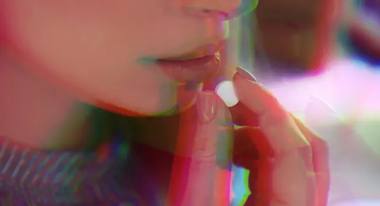 A close-up of a woman’s lips as she holds a small pill, with a colorful, blurred effect surrounding her face.