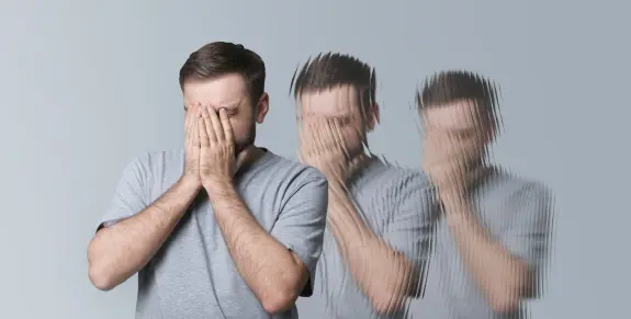 A man in a gray shirt covers his face with his hands, with blurred figures of himself in the background.