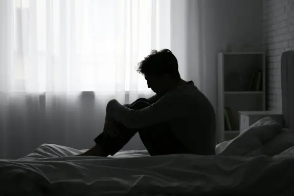 A silhouette of a man sitting on a bed, hugging his knees, in a softly lit, minimalistic room.