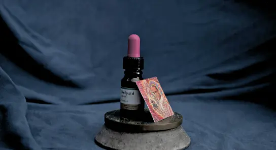 A dropper bottle with a pink cap beside a colorful psychedelic art piece, set on a circular concrete base against a dark b...