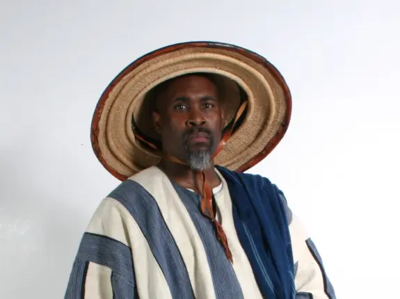 Man wearing a traditional straw hat and a patterned garment, standing against a plain white background.