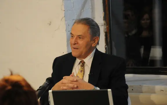Elderly man in a suit sits at a table, hands clasped, speaking with an audience in a dimly lit room.