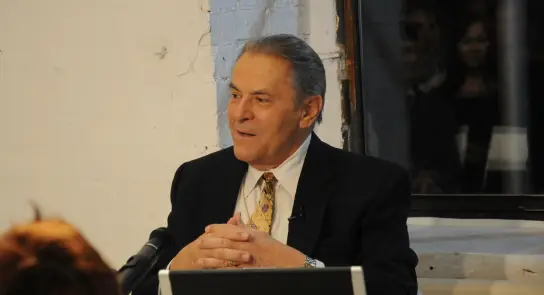 Elderly man in a suit sits at a table, hands clasped, speaking with an audience in a dimly lit room.