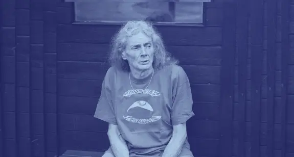 Elderly person with long gray hair, wearing a graphic t-shirt, seated against a dark wooden backdrop.