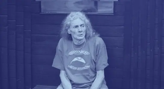 Elderly person with long gray hair, wearing a graphic t-shirt, seated against a dark wooden backdrop.
