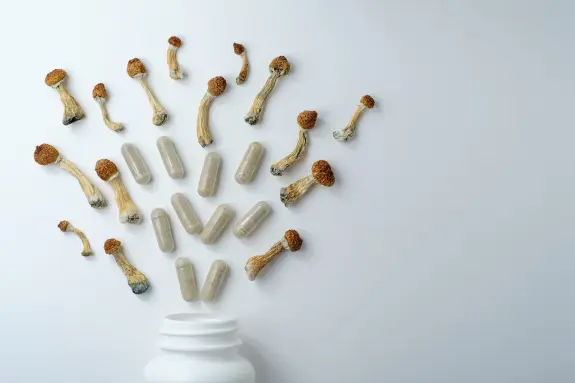 Dried psilocybin mushrooms and transparent capsules scattered around a white bottle on a light background.