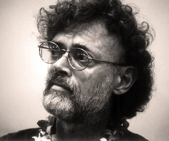 A man with curly hair and glasses gazes thoughtfully, wearing a floral lei around his neck.