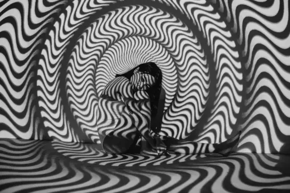 A figure sits against a swirling black and white patterned backdrop, creating a hypnotic visual effect.