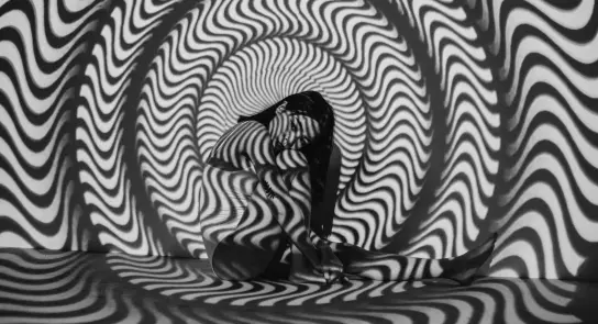 A figure sits against a swirling black and white patterned backdrop, creating a hypnotic visual effect.