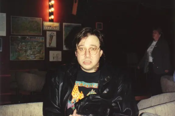 Man with glasses and a leather jacket, looking surprised, seated in a dimly lit room with artwork on the walls.