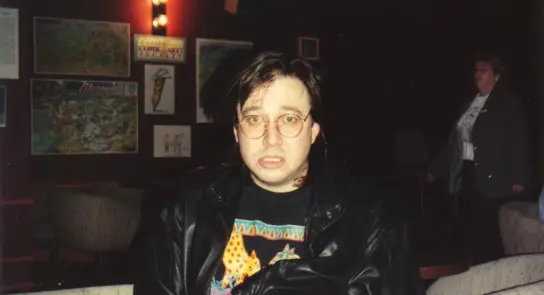 Man with glasses and a leather jacket, looking surprised, seated in a dimly lit room with artwork on the walls.