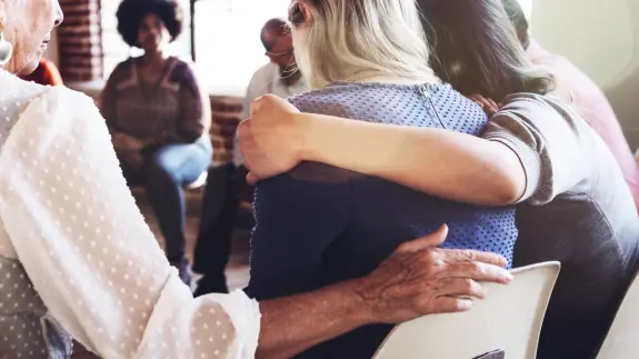 Two women embrace in a supportive group setting, with others seated in the background, fostering a sense of community.